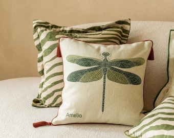 May include: A square throw pillow featuring a green dragonfly graphic and the name Amelia printed in green. The pillow has dark red piping and tassels at the corners. Behind it sits a second pillow with a green and white zebra stripe pattern.