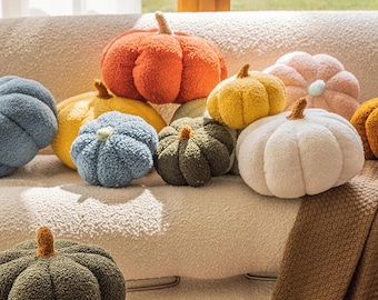 May include: A collection of plush pumpkin-shaped pillows in various colors, including orange, yellow, green, blue, and white. Each pillow has a stem and is arranged on a beige couch, creating a cozy, autumnal theme.