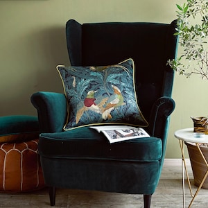 Può includere: Una poltrona in velluto verde acqua con un cuscino quadrato decorativo con un disegno di uccelli e fogliame. Il cuscino ha un bordo dorato. Un secondo cuscino è visibile sulla sedia. Un tavolino con piano in marmo sulla destra.