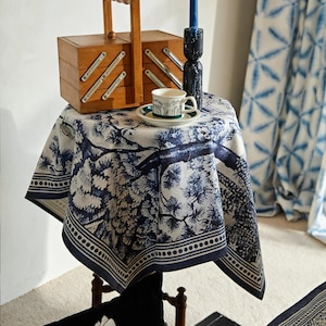 May include: A wooden sewing box with a handle rests on a small table draped with a blue and white patterned tablecloth. A teacup and saucer with a green and white design are on the table, along with a black candle holder and a blue candle.