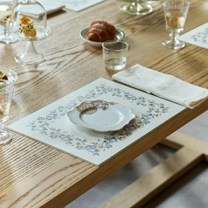 May include: A rectangular placemat featuring a white background with a border of blue flowers and green leaves. A small, white, scalloped-edge saucer with a brown floral design sits in the center of the placemat, next to a folded white napkin.