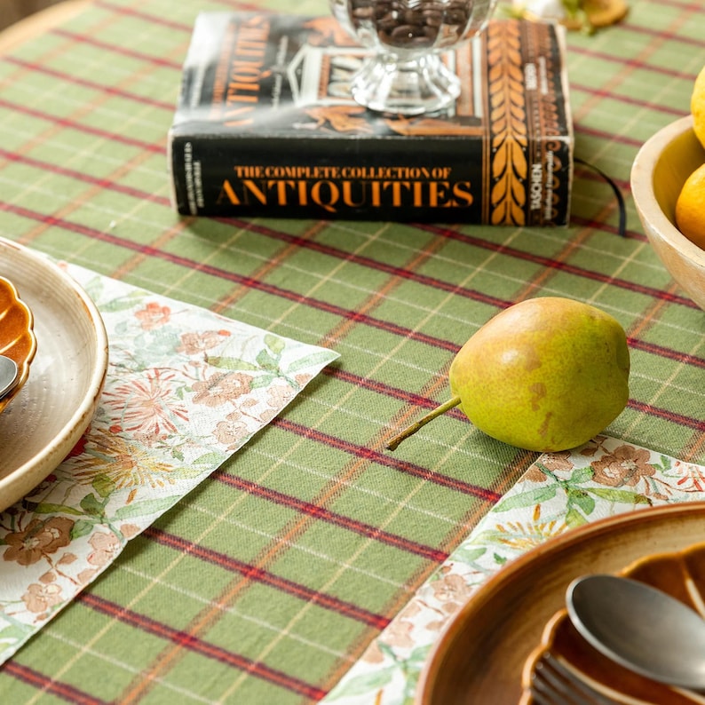 May include: A still-life scene featuring a green and red plaid tablecloth. A book titled "The Complete Collection of Antiquities" sits atop the cloth, along with a pear, a bowl of chocolates, and a plate. The tablecloth has floral placemats.