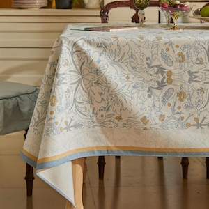 以下が含まれることがあります： クリーム色のテーブルクロス。グレーとイエローの繊細な花と鳥の模様が特徴。縁には青と黄色のストライプ。木製のテーブルに掛けられており、ダイニングや装飾的な場面を連想させる。