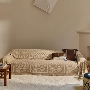 Puede incluir: Un sofá beige cubierto con una manta estampada con formas geométricas y motivos de árboles. Dos cojines rectangulares están colocados sobre el sofá, uno marrón con borlas azules y otro blanco con una sección superior de cuero marrón.