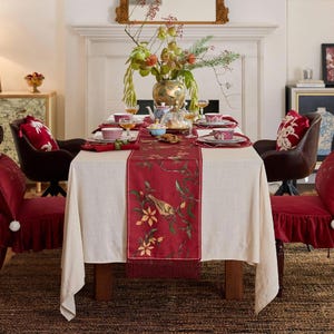 May include: A dining table set for a meal, featuring a cream-colored tablecloth and a red runner with a bird and floral design. The table is set with plates, cups, and a teapot. Two chairs with red cushions are on either side of the table.