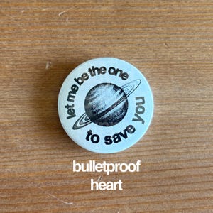 May include: A round, light blue button with the text "Let me be the one to save you" encircling a black and white illustration of a planet with rings. Below the planet is the text "bulletproof heart".