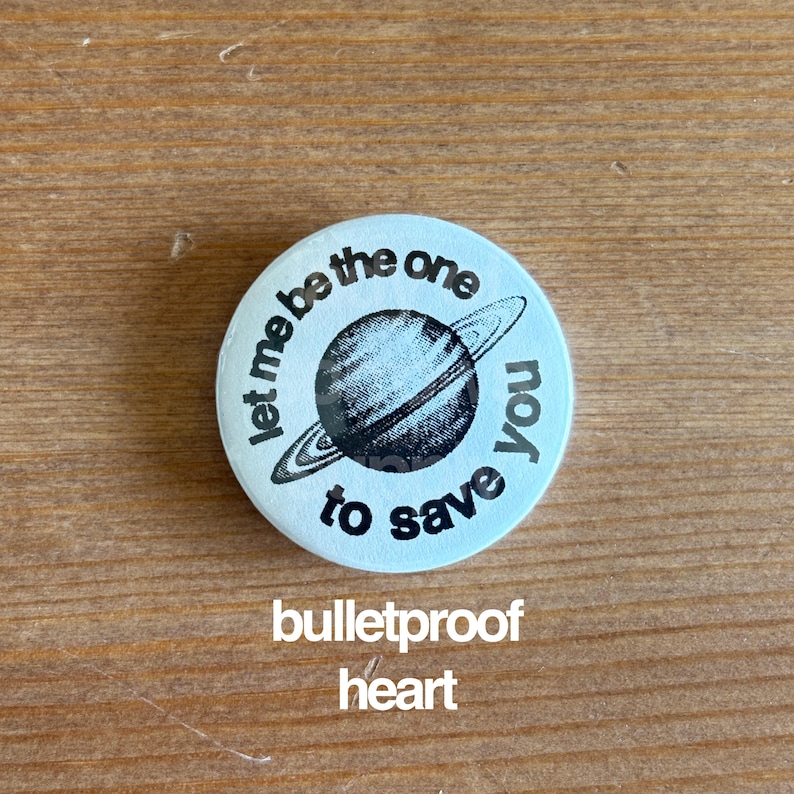 May include: A round, light blue button with the text "Let me be the one to save you" encircling a black and white illustration of a planet with rings. Below the planet is the text "bulletproof heart".