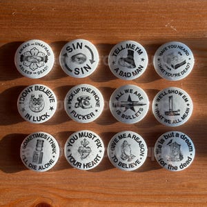 May include: A collection of twelve white round buttons with black text and illustrations. The buttons feature phrases like "Awake and Unafraid," "Sin," "Tell Me I'm a Bad Man," and "I Don't Believe in Luck." The buttons are arranged on a wooden surface.