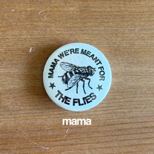 May include: A round, light blue button with the text "MAMA WE'RE MEANT FOR THE FLIES" encircling a detailed illustration of a fly. The word "mama" is printed at the bottom. Two stars flank the fly illustration.