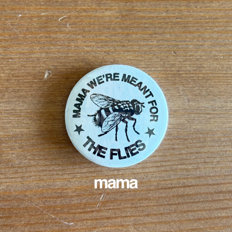May include: A round, light blue button with the text "MAMA WE'RE MEANT FOR THE FLIES" encircling a detailed illustration of a fly. The word "mama" is printed at the bottom. Two stars flank the fly illustration.