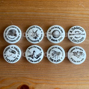 May include: A collection of eight white round buttons with various black and white graphics and text. The buttons feature phrases like "Never Coming Home," "Think Happy Thoughts," and "A Dagger."