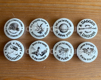 Puede incluir: Una colección de ocho botones redondos blancos con varios gráficos y texto en blanco y negro. Los botones presentan frases como "Never Coming Home", "Think Happy Thoughts" y "A Dagger".