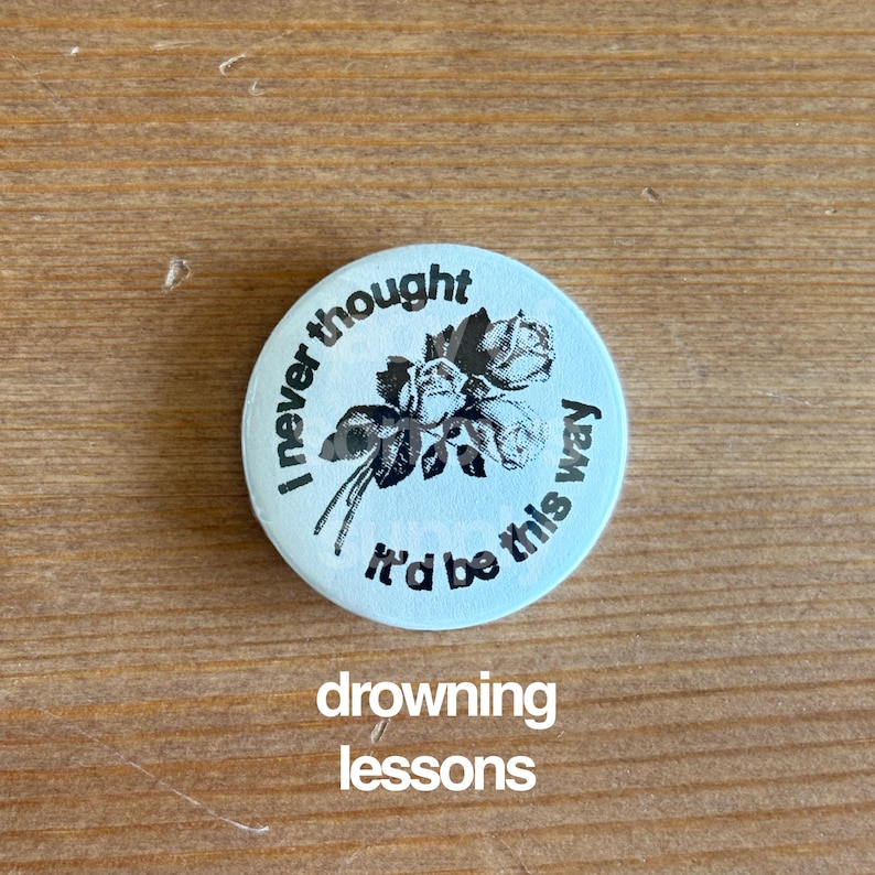 May include: A light blue button with the text "I never thought it'd be this way" encircling a black and white floral design. The words "drowning lessons" are printed below the button.