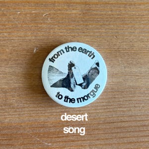 May include: A round, light blue button with the text "from the earth to the morgue" in a circular pattern. The button features a black and white graphic of feet with a toe tag. Below the button, the words "desert song" are printed.