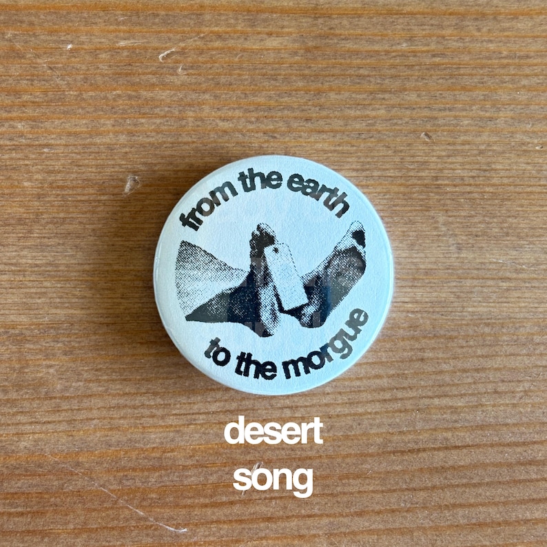 May include: A round, light blue button with the text "from the earth to the morgue" in a circular pattern. The button features a black and white graphic of feet with a toe tag. Below the button, the words "desert song" are printed.