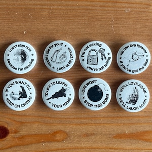 Może przedstawiać: Osiem białych, okrągłych guzików z czarnym tekstem i ilustracjami. Guziki zawierają frazy takie jak "I can't stop now because I'm dancing" i "Love won't stop this bomb". Ułożone są na drewnianej powierzchni.