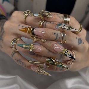 May include: An array of gold rings and nail art. The nails are long and pointed, with a variety of designs including gold accents, gems, and a French manicure style. The rings feature intricate details and various gemstones.