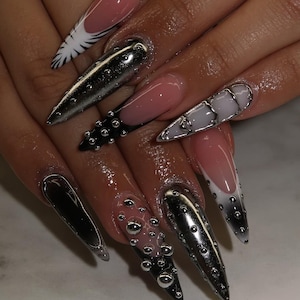 May include: A set of long, pointed acrylic nails in various designs. Some nails are chrome, others have a French tip with white and black accents, and some feature clear embellishments.