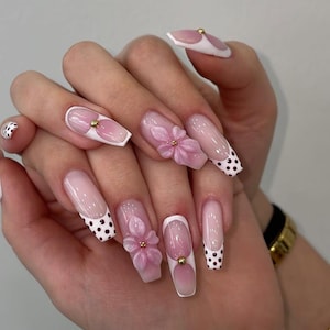 May include: A set of long, square-shaped acrylic nails. The nails feature a gradient pink and white design, with some adorned with 3D pink flowers and gold accents. Some nails have a white tip with black polka dots.