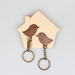 May include: A wooden key holder in the shape of a house, with two bird silhouettes in shades of brown and tan. It has two brass-coloured key rings suspended by chains, ideal for key organisation. The background is plain white.