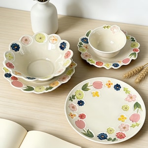 Może przedstawiać: Kolekcja kremowej ceramiki stołowej z kwiatowymi wzorami. Zestaw zawiera dużą miskę, mniejszą miskę ze spodkiem i talerz obiadowy. Kwiatowe wzory mają różowe, niebieskie, żółte i zielone akcenty, tworząc wesołą estetykę.