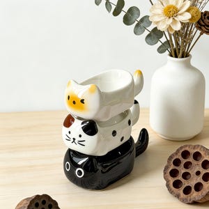 Puede incluir: Cuencos apilados con forma de gato de cerámica en blanco, negro y gris. El cuenco superior es blanco con detalles amarillos, el del medio es gris con lunares negros y el inferior es negro. Un jarrón blanco con flores secas a la derecha.