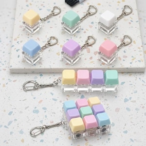 May include: A collection of keycap keychains in various pastel colors including yellow, mint green, white, blue, purple, and pink. The keychains feature single keycaps, a row of four keycaps, and a three-by-three grid of nine keycaps.