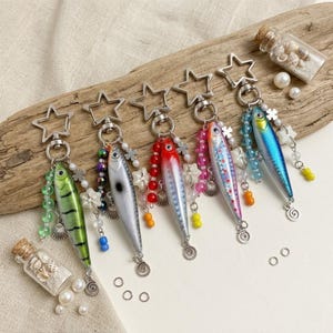 May include: Six colorful fish-shaped keychains with star-shaped clasps and beaded accents. Each keychain features a different color scheme, including green, silver, red, pink, and blue. Small glass bottles filled with shells and pearls are also present.