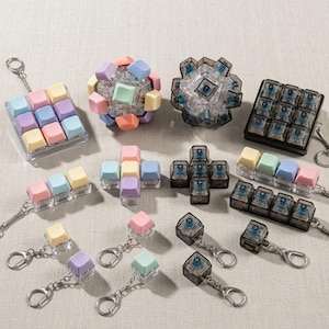 May include: A collection of mechanical keyboard switch keychains in various configurations, including single, triple, and nine-key pads. The keycaps feature pastel colors like pink, blue, yellow, and purple, alongside transparent black housings with blue switches.