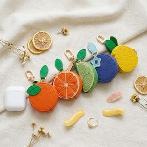 May include: A collection of round, textured, fruit-shaped cases with gold-tone metal keychains. The designs include an orange, a lime wedge, a blue berry with a star charm, and a yellow pineapple. A white charging case is positioned to the left.