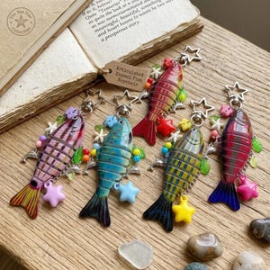 May include: Articulated enamel fish keychains in various colors, including pink, blue, red, and yellow. Each keychain features a fish design with star and bead accents. The keychains are displayed on a wooden surface.