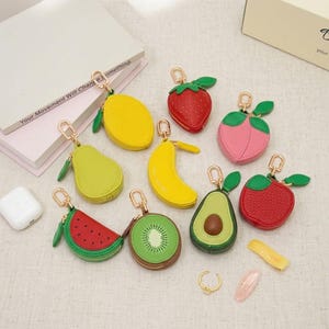 Pode incluir: Uma coleção de porta-moedas em forma de fruta com porta-chaves de metal dourado. O conjunto inclui um limão amarelo, um morango vermelho, um pêssego rosa, uma pera verde, uma banana amarela, uma melancia, um kiwi, um abacate e uma maçã vermelha.