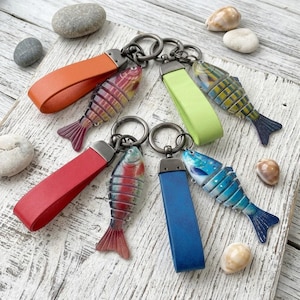 May include: A collection of fish-shaped keychains with colorful leather straps in orange, red, green, and blue. The fish are detailed with various colors and patterns, attached to silver key rings.