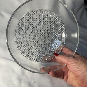 May include: Clear glass plate with a textured, patterned center. The plate has a wide, smooth rim and is held by a hand with red nail polish. The center of the plate features a repeating diamond and circle design.