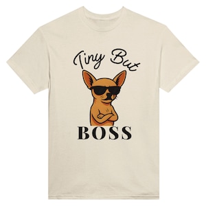 Könnte beinhalten: Cremefarbenes T-Shirt mit einem Cartoon-Chihuahua, der eine Sonnenbrille trägt und die Arme verschränkt hat. Der Text "Tiny But Boss" befindet sich über und unter der Hundeillustration. Das Shirt hat einen Rundhalsausschnitt und kurze Ärmel.