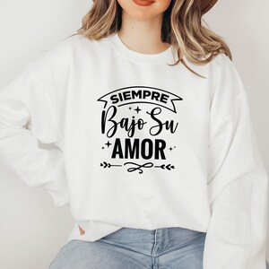 Może przedstawiać: Biała bluza z hiszpańskim napisem "SIEMPRE Bajo Su AMOR" w czarnym skrypcie. Projekt zawiera gwiazdy i elementy dekoracyjne. Bluza jest w stylu casual.