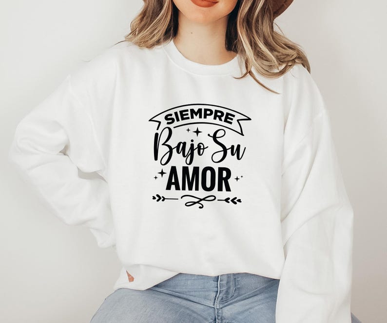 Może przedstawiać: Biała bluza z hiszpańskim napisem "SIEMPRE Bajo Su AMOR" w czarnym skrypcie. Projekt zawiera gwiazdy i elementy dekoracyjne. Bluza jest w stylu casual.