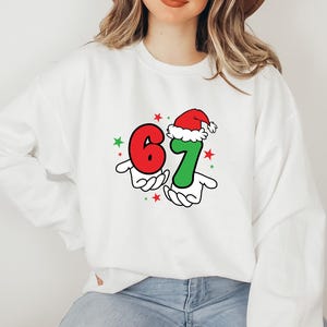May include: White sweatshirt featuring the numbers "67" in red and green, held by cartoon hands. The number "7" is topped with a Santa hat. Red and green stars are scattered around the numbers, creating a festive holiday design.