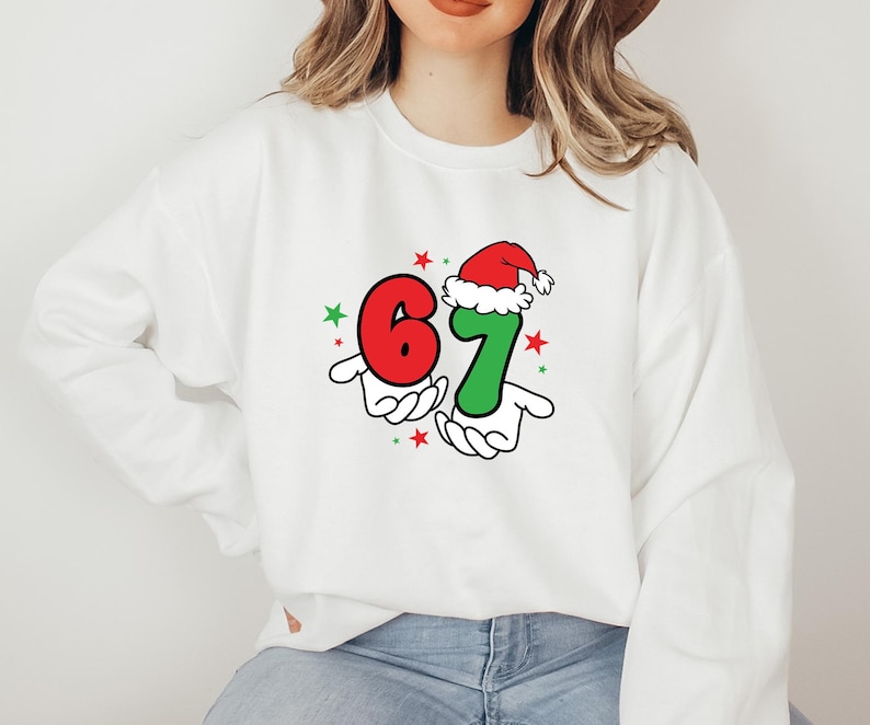 May include: White sweatshirt featuring the numbers "67" in red and green, held by cartoon hands. The number "7" is topped with a Santa hat. Red and green stars are scattered around the numbers, creating a festive holiday design.