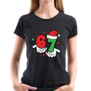 May include: Black t-shirt featuring the numbers 6 and 7 in red and green, held by cartoon hands. The number 7 is wearing a Santa hat. Red and green stars are scattered around the numbers, creating a festive design.