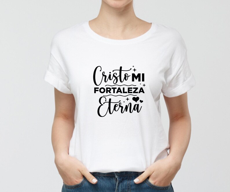 Może przedstawiać: Biała koszulka z napisem "Cristo MI Fortaleza Eterna" w czarnym skrypcie. Koszulka ma kr&oacute;tkie rękawy i okrągły dekolt. Tekst zawiera ozdobne gwiazdki i serca.