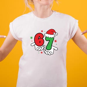 May include: White t-shirt featuring a festive graphic with the numbers 6 and 7, held by cartoon hands. The number 7 is wearing a Santa hat. Red and green stars complete the design. The shirt is on a person with a yellow background.