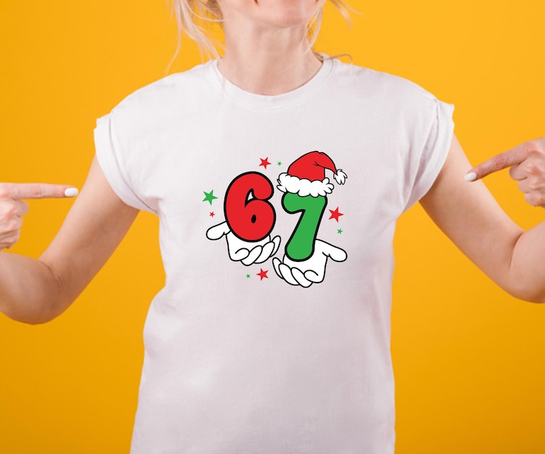 May include: White t-shirt featuring a festive graphic with the numbers 6 and 7, held by cartoon hands. The number 7 is wearing a Santa hat. Red and green stars complete the design. The shirt is on a person with a yellow background.