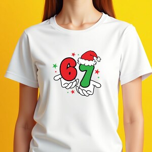 May include: White t-shirt featuring a festive design with the numbers "6" and "7" in red and green, respectively. The number "7" is adorned with a Santa hat. The design includes red and green stars, and the numbers are held by cartoon hands.
