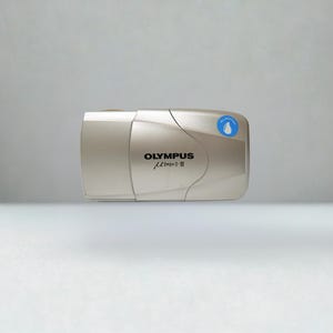 May include: A champagne-coloured Olympus mju: II 35mm film camera with a sliding lens cover. The front features the black text OLYMPUS and the model name, along with a small circular blue sticker with a white water drop icon and the text ALL WEATHER.