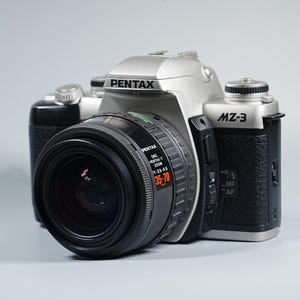 Könnte beinhalten: Eine silberne und schwarze Pentax MZ-3 Kamera mit einem 35-70mm Objektiv. Das Kameragehäuse trägt das Pentax-Logo und den Modellnamen MZ-3. Das Objektiv trägt die Aufschrift "Pentax F Zoom 1:3.5-4.5 35-70".