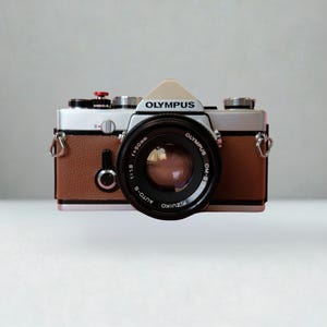 May include: An Olympus film camera with a silver top, brown textured body, and a black lens. The lens is labelled Olympus OM-SYSTEM F.ZUIKO AUTO-S 1:1.8 f=50mm. The camera features a red shutter button, silver dials, and metal strap lugs on the sides.