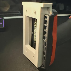 Puede incluir: Un marco gris claro impreso en 3D con una abertura rectangular, que sostiene un objeto negro con texto blanco. Una herramienta eléctrica negra y roja es parcialmente visible en el lado derecho del marco.