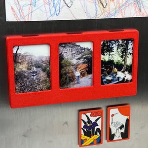 May include: A rectangular red magnetic frame with three vertical slots holding photographs of outdoor scenes. Below the frame are two small rectangular magnets, one featuring a purple iris flower and the other a white crane illustration.