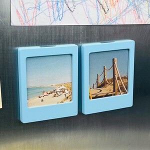 May include: Two light blue square magnetic photo frames attached to a metallic surface. The left frame displays a beach scene with sand and water, while the right frame shows a wooden boardwalk with rope railings leading through grassy dunes.